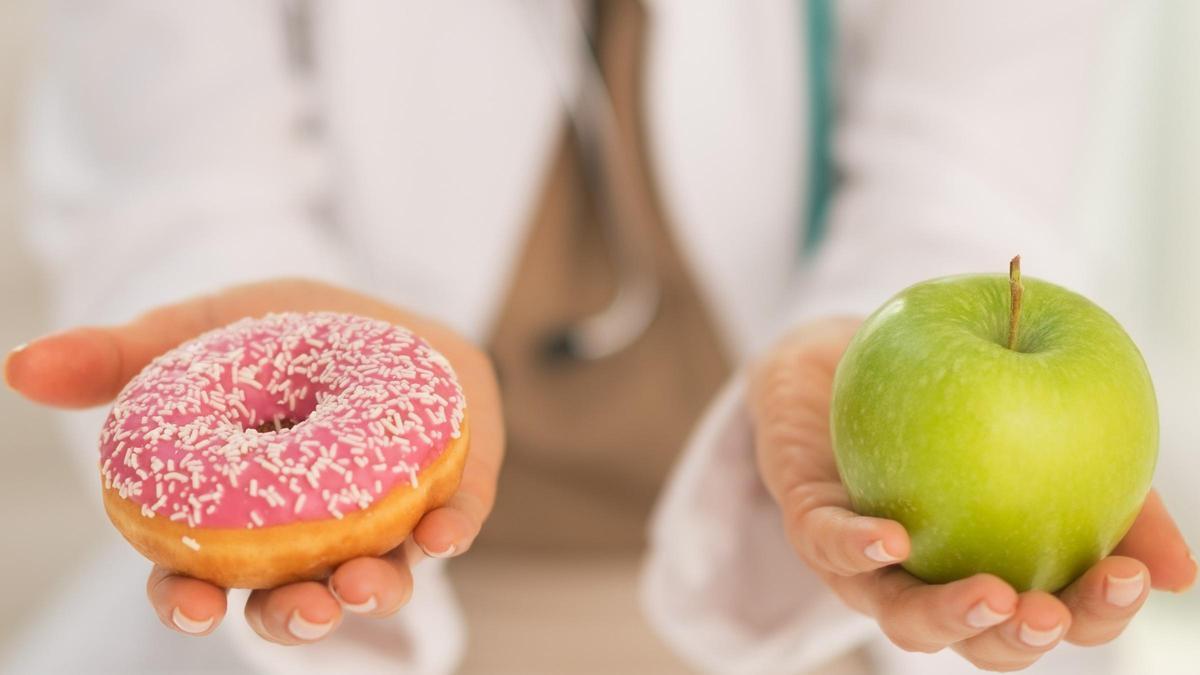 Una doctora sostiene un donut o rosquilla en una mano y una manzana en la otra Una doctora sostiene un donut o rosquilla en una mano y una manzana en la otra