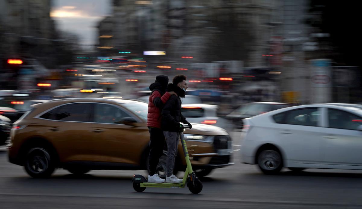 Dos jóvenes desafían al tráfico con un patín eléctrico en el centro de Madrid.