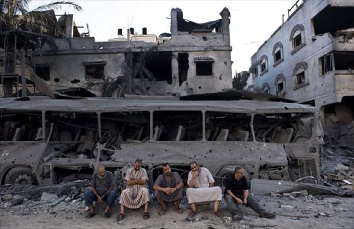 Cinco palestinos de Gaza sentados frente a un autobús y un edificio destruidos, en el 2014, por ataques aéreos israelís. Cinco palestinos de Gaza sentados frente a un autobús y un edificio destruidos, en el 2014, por ataques aéreos israelís.