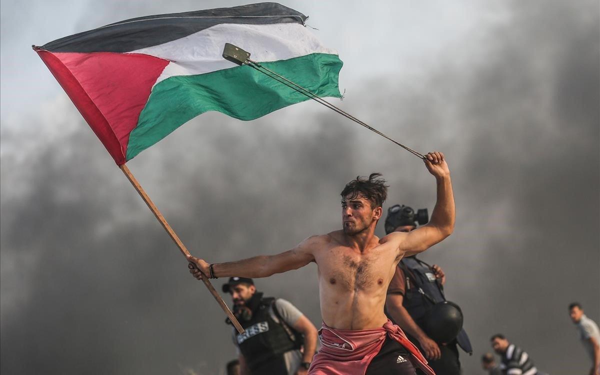 Un joven palestino tira piedras con una honda mientras con la otra mano lleva la bandera palestina.