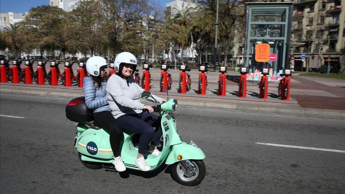Dos usuarias de moto compartida pasan por delante de una parada del Bicing. Dos usuarias de moto compartida pasan por delante de una parada del Bicing.
