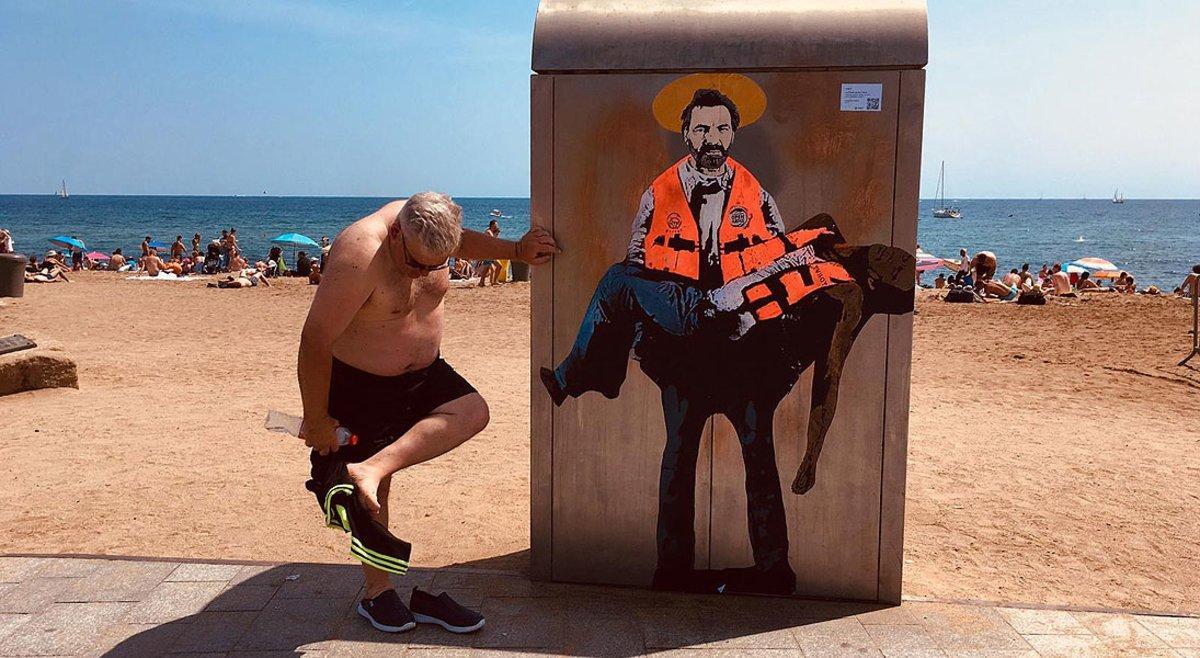 Un bañista se limpia la arena de los pies junto al dibujo de Òscar Camps pintado por Tvboy en la Barceloneta.