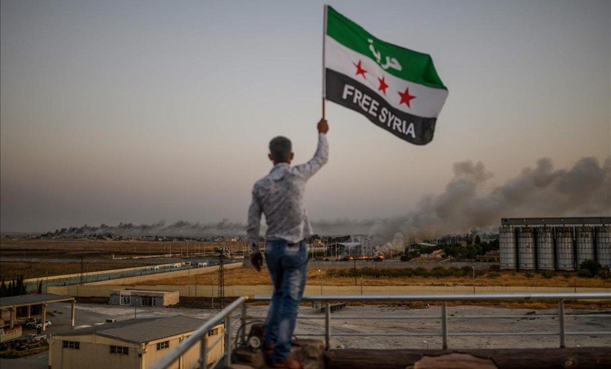 Un hombre sostiene una bandera en la que pide la liberación de Siria mientras las columnas de humo se elevan en las localidades fronterizas bombardeadas este domingo.
