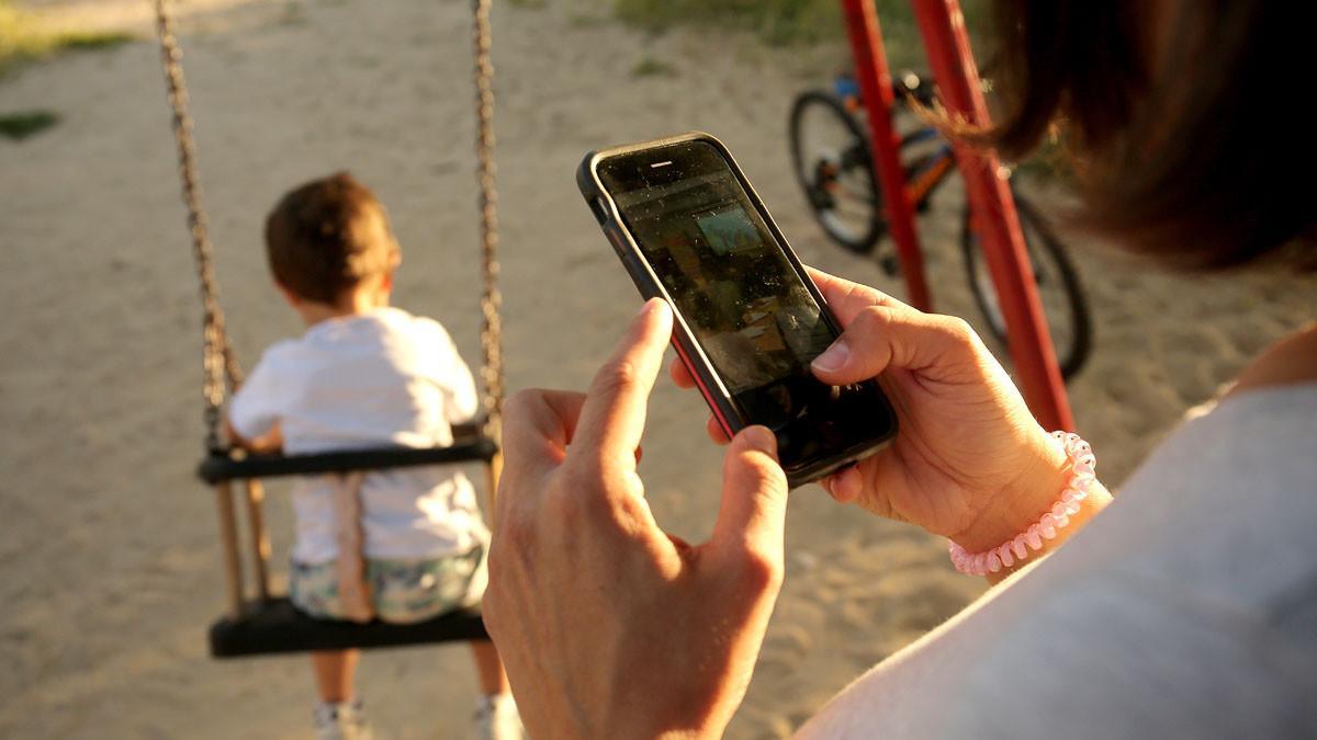 Una madre mira su teléfono móvil mientras su hija juega en el parque.