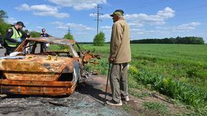Un anciano observa cómo oficiales de policía examinan un coche quemado tras recibir el impacto de munición rusa, en la localidad ucraniana de Mala Rogan.