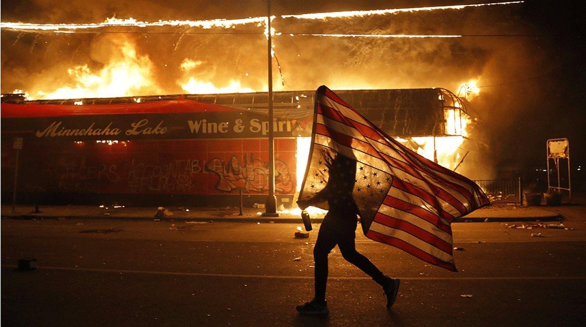 Un manifestante porta una bandera estadounidense al revés, junto a un edificio en llamas, en Minneápolis el jueves 28 de mayo. Un manifestante porta una bandera estadounidense al revés, junto a un edificio en llamas, en Minneápolis el jueves 28 de mayo.