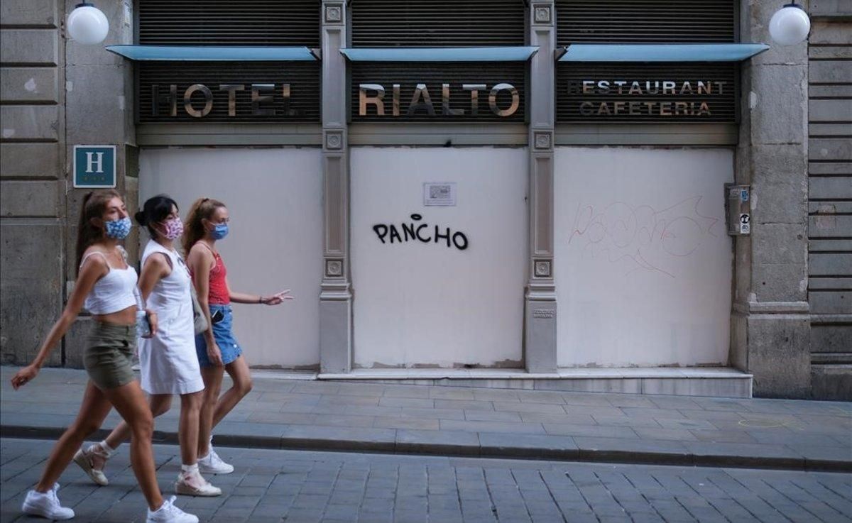 Jóvenes con mascarilla pasan por delante de la entrada de un hotel cerrado, en Barcelona. Jóvenes con mascarilla pasan por delante de la entrada de un hotel cerrado, en Barcelona.