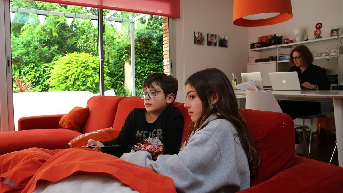 Irene y Mauro Bagen Guillem, de 12 y 9 años, en su casa en Barcelona mirando la tele. Al fondo, su madre teletrabajando. Irene y Mauro Bagen Guillem, de 12 y 9 años, en su casa en Barcelona mirando la tele. Al fondo, su madre teletrabajando.