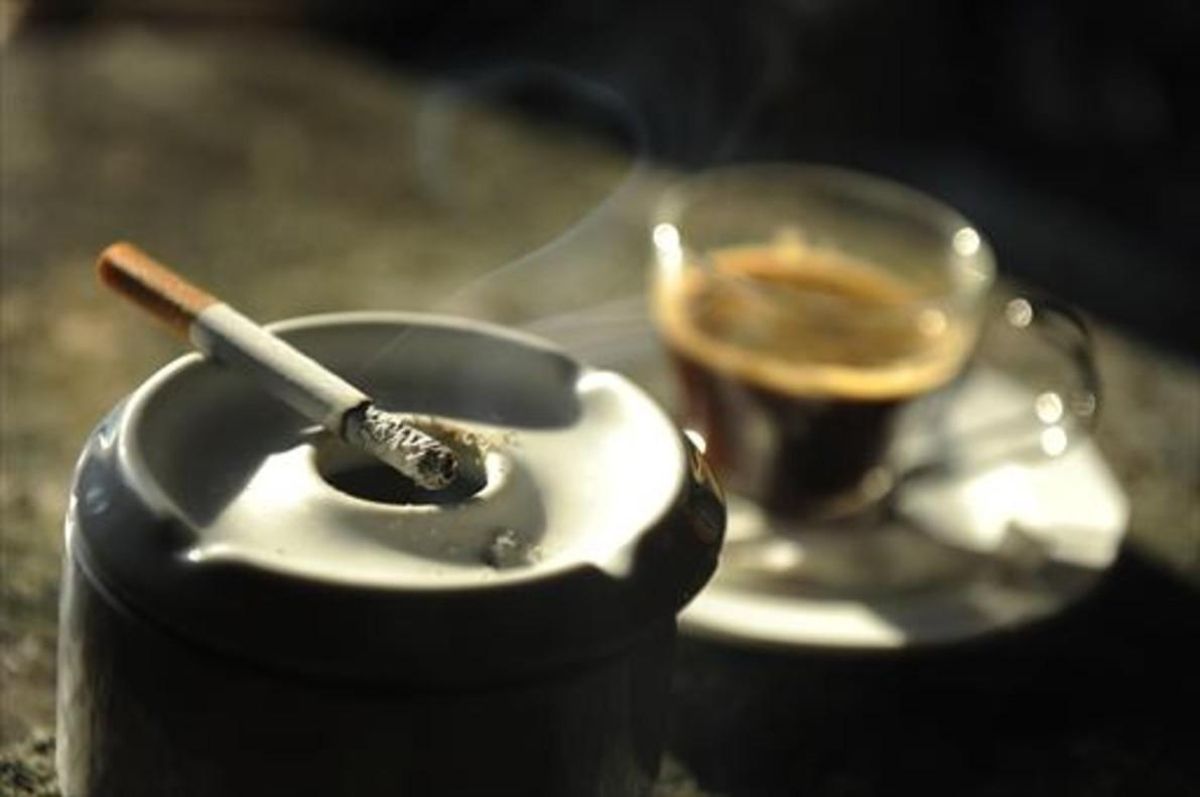 Un cigarrillo humea junto a una taza de café en un bar. Un cigarrillo humea junto a una taza de café en un bar.