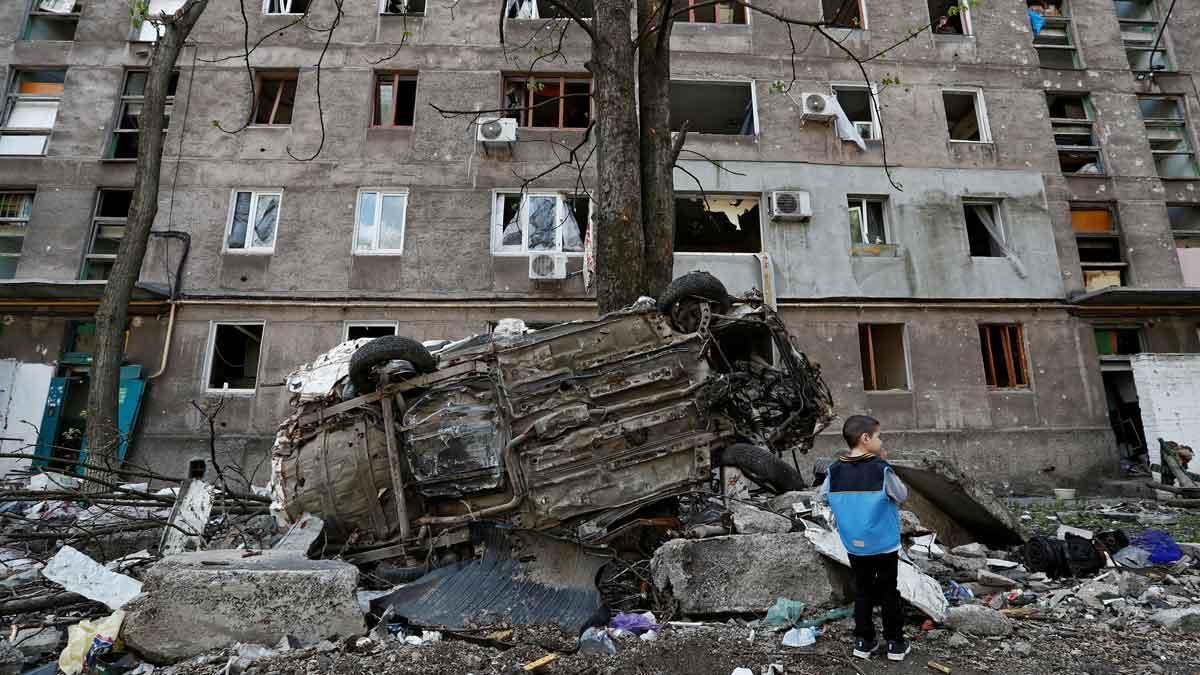 Un niño junto a un coche destrozado y un edificio de viviendas dañado por los ataques en la ciudad de Mariúpol.